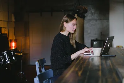 Side view of young woman working on netbook sitting at table with cup of hot beverage in kitchen