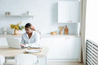 A black woman types on her laptop in a bright, modern kitchen, embodying work-from-home lifestyle.