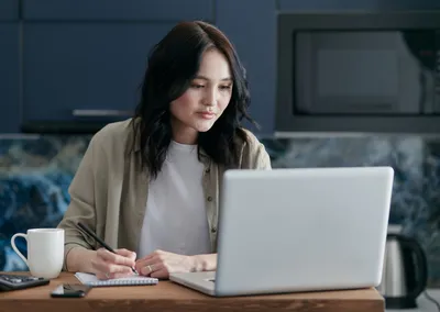 A focused woman takes notes while working on her laptop in a modern kitchen with coffee.