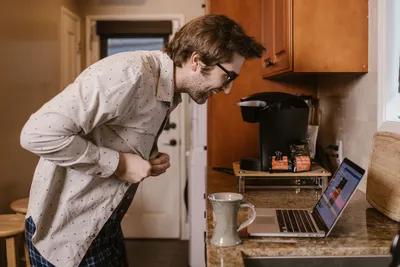 Casual man buttoning shirt in home kitchen while engaging in remote work on laptop.