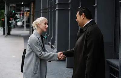 Side view of cheerful stylish diverse coworkers in elegant coats shaking hands and looking at each other on street